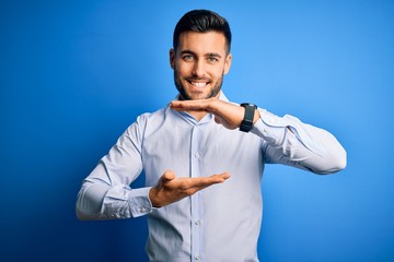 Young handsome man wearing elegant shirt standing over isolated blue background gesturing with hands showing big and large size sign, measure symbol. Smiling looking at the camera. Measuring concept.