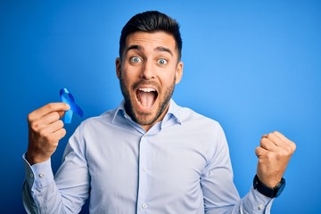 Young handsome man holding blue ribbon as prostate campaing support over blue background screaming proud and celebrating victory and success very excited, cheering emotion