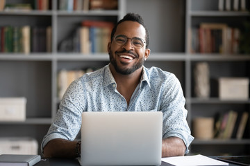 Portrait of smiling African American man in glasses sit at desk in office working on laptop, happy...