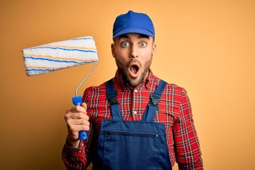 Young handsome painter man painting wall using roller over isolated yellow background scared in...