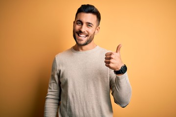 Young handsome man wearing casual sweater standing over isolated yellow background doing happy thumbs up gesture with hand. Approving expression looking at the camera showing success. - obrazy, fototapety, plakaty