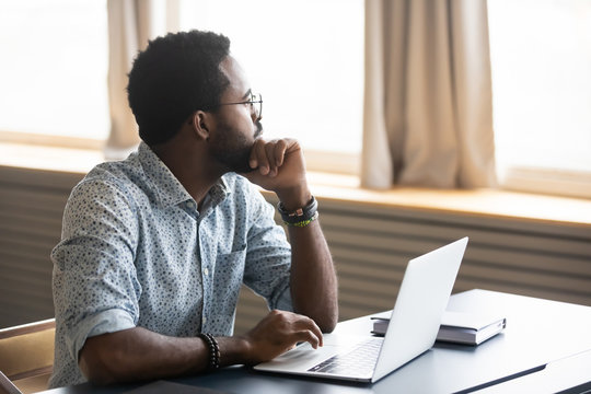 Thoughtful african American male employee in glasses sit at desk distracted from computer work pondering, pensive biracial man look in distance thinking or planning, business vision concept