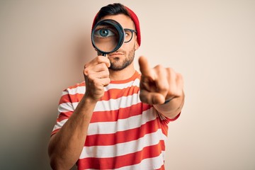 Young detective man looking through magnifying glass over isolated background pointing with finger to the camera and to you, hand sign, positive and confident gesture from the front