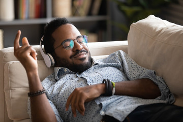 Happy biracial millennial man in wireless headphones relax on couch at home listen to music,...
