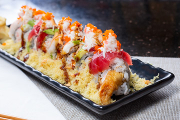 A closeup view of a decorated sushi roll with tuna, avocado and shrimp tempura, in a restaurant or kitchen setting.