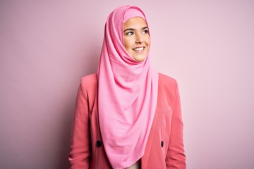 Young beautiful girl wearing muslim hijab standing over isolated pink background looking away to side with smile on face, natural expression. Laughing confident.