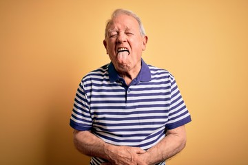 Grey haired senior man wearing casual navy striped t-shirt standing over yellow background sticking tongue out happy with funny expression. Emotion concept.