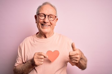 Senior grey haired man holding heart shape paper over pink background happy with big smile doing ok...