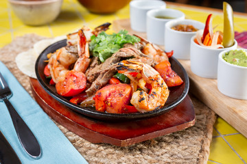 A view of a sizzling skillet plate of surf and turf style fajitas, featuring several condiments and dips on the side, in a restaurant or kitchen setting.