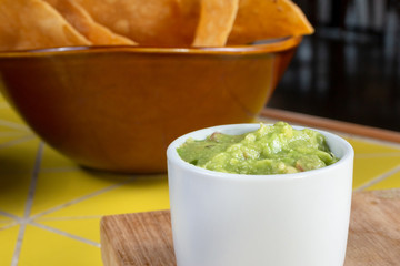 A closeup view of a cup of guacamole, with a background bowl of corn tortilla chips, in a restaurant or kitchen setting.