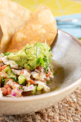 A closeup view of a bowl of ceviche, garnished with guacamole and an array of deep fried tortillas, in a restaurant or kitchen setting.
