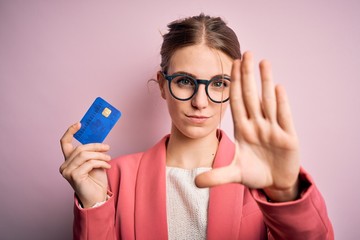 Young beautiful redhead woman holding credit card over isolated pink background with open hand doing stop sign with serious and confident expression, defense gesture