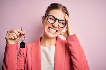 Young beautiful redhead house agent woman holding home key over pink bakcground stressed with hand...