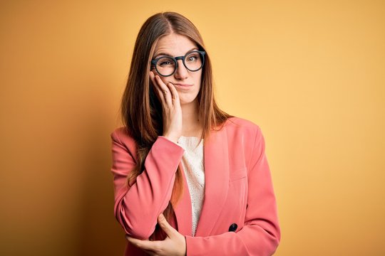 Young beautiful redhead woman wearing jacket and glasses over isolated yellow background thinking looking tired and bored with depression problems with crossed arms.