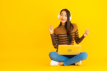 Beautiful portrait young asian woman sit on the floor with laptop and headphone on yellow background