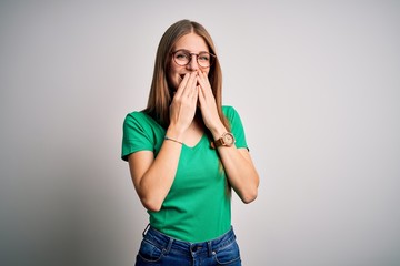 Young beautiful redhead woman wearing casual green t-shirt and glasses over white background laughing and embarrassed giggle covering mouth with hands, gossip and scandal concept