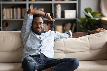 Happy biracial millennial man sit relax on comfortable sofa in living room watch TV, smiling calm...