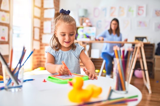 Caucasian girl kid playing and learning at playschool with female teacher. Mother and daughter at playroom around toys drawing on magnetic blackboard