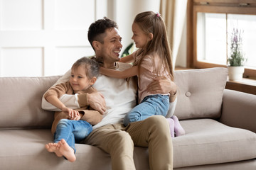 Smiling young father sitting on comfortable couch, playing with small cute kids daughters in living...