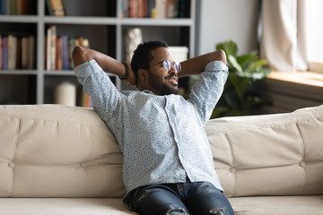 Happy smiling African American young man sit on sofa at home look in distance thinking pondering,...