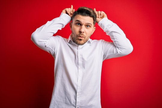 Young business man with blue eyes wearing elegant shirt standing over red isolated background doing funny gesture with finger over head as bull horns