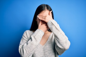 Young beautiful asian woman wearing casual sweater standing over blue isolated background Covering eyes and mouth with hands, surprised and shocked. Hiding emotion