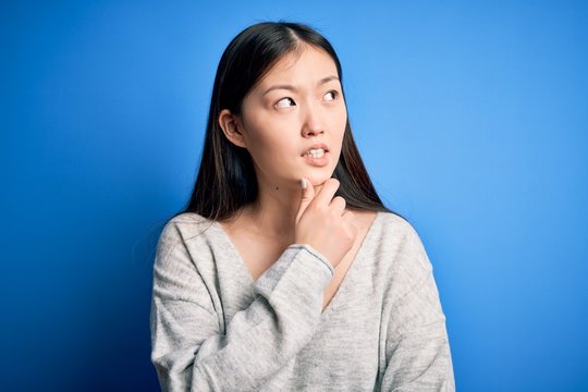 Young beautiful asian woman wearing casual sweater standing over blue isolated background Thinking worried about a question, concerned and nervous with hand on chin