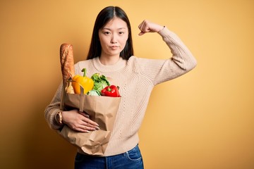 Young asian woman holding paper bag of fresh healthy groceries over yellow isolated background Strong person showing arm muscle, confident and proud of power