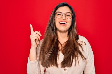 Young hispanic smart woman wearing glasses standing over red isolated background showing and pointing up with finger number one while smiling confident and happy.