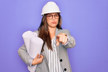 Young hispanic architect woman wearing builder hat and holding blueprints over yellow background pointing with finger to the camera and to you, hand sign, positive and confident gesture from the front