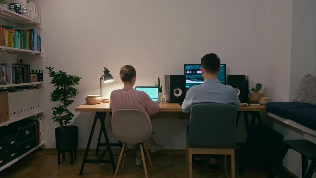 Couple Of Two Remote Workers Are Working From Home In Evening Or Night. Editor, Bloggers Or Freelancers Sit At Computer And Laptop In Cozy Room. Family, Partners Or Spouses Work Together. 4K Wide Shot