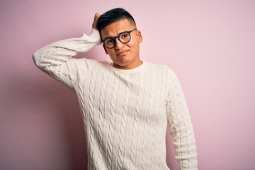 Young handsome latin man wearing white casual sweater and glasses over pink background confuse and wonder about question. Uncertain with doubt, thinking with hand on head. Pensive concept.