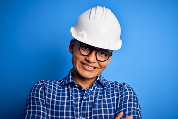 Young handsome engineer latin man wearing safety helmet over isolated blue background happy face smiling with crossed arms looking at the camera. Positive person.