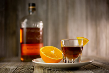 A view of a shot of whiskey with a half orange on a plate, and a whiskey bottle in the background.