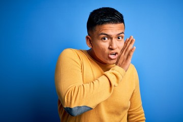 Young handsome latin man wearing yellow casual sweater over isolated blue background hand on mouth telling secret rumor, whispering malicious talk conversation
