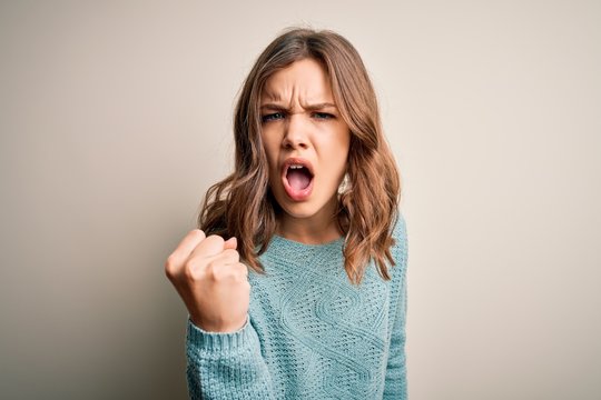 Young blonde girl wearing casual blue winter sweater over isolated background angry and mad raising fist frustrated and furious while shouting with anger. Rage and aggressive concept.