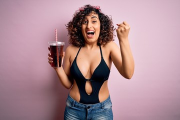 Beautiful arab woman on vacation wearing swimsuit drinking cola refreshment using straw screaming proud and celebrating victory and success very excited, cheering emotion