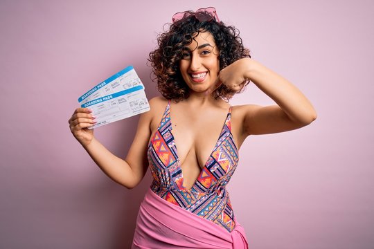 Young beautiful tourist arab woman on vacation wearing swimsuit holding boarding pass airline with surprise face pointing finger to himself