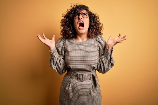Beautiful arab business woman wearing dress and glasses standing over yellow background crazy and mad shouting and yelling with aggressive expression and arms raised. Frustration concept.