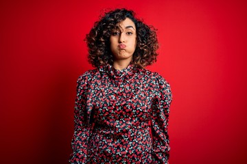 Young beautiful curly arab woman wearing casual floral dress standing over red background puffing cheeks with funny face. Mouth inflated with air, crazy expression.