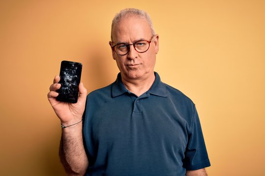 Middle age man holding broken smartphone showing cracked screen over yellow background with a confident expression on smart face thinking serious