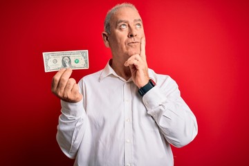 Middle age hoary man holding one dollar banknote over isolated red background serious face thinking about question, very confused idea
