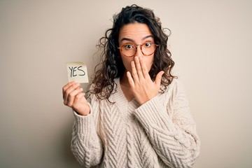 Young beautiful woman with curly hair holding reminder paper with yes word message cover mouth with...