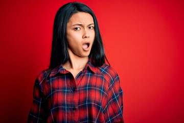 Young beautiful chinese woman wearing casual shirt over isolated red background In shock face,...