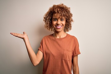 Beautiful african american woman with curly hair wearing casual t-shirt over white background smiling cheerful presenting and pointing with palm of hand looking at the camera.