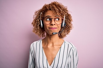 African american curly call center agent woman working using headset over pink background smiling looking to the side and staring away thinking.