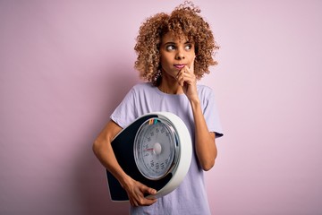 African american slim woman with curly hair holding scale over isolated pink background serious face thinking about question, very confused idea