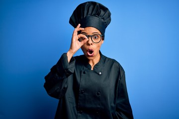 Young african american chef woman wearing cooker uniform and hat over blue background doing ok gesture shocked with surprised face, eye looking through fingers. Unbelieving expression.