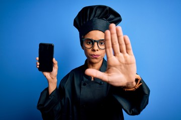 African american baker woman wearing cooker uniform holding smartphone showing screen with open hand doing stop sign with serious and confident expression, defense gesture