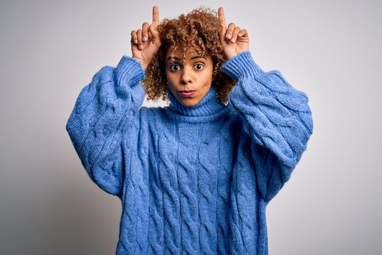 Young beautiful african american woman wearing turtleneck sweater over white background doing funny gesture with finger over head as bull horns